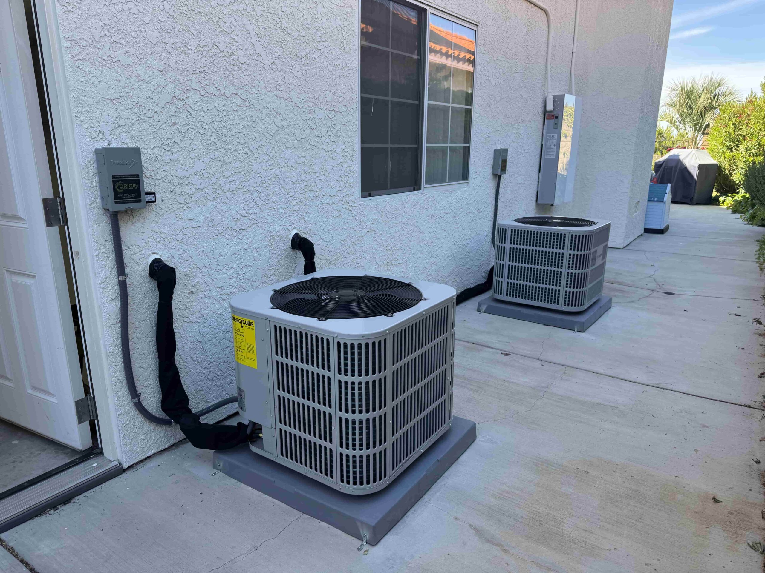 Two newly installed residential air conditioning condenser units mounted on concrete pads outside a stucco home in Palmdale, serviced by Origin Heating & Air
