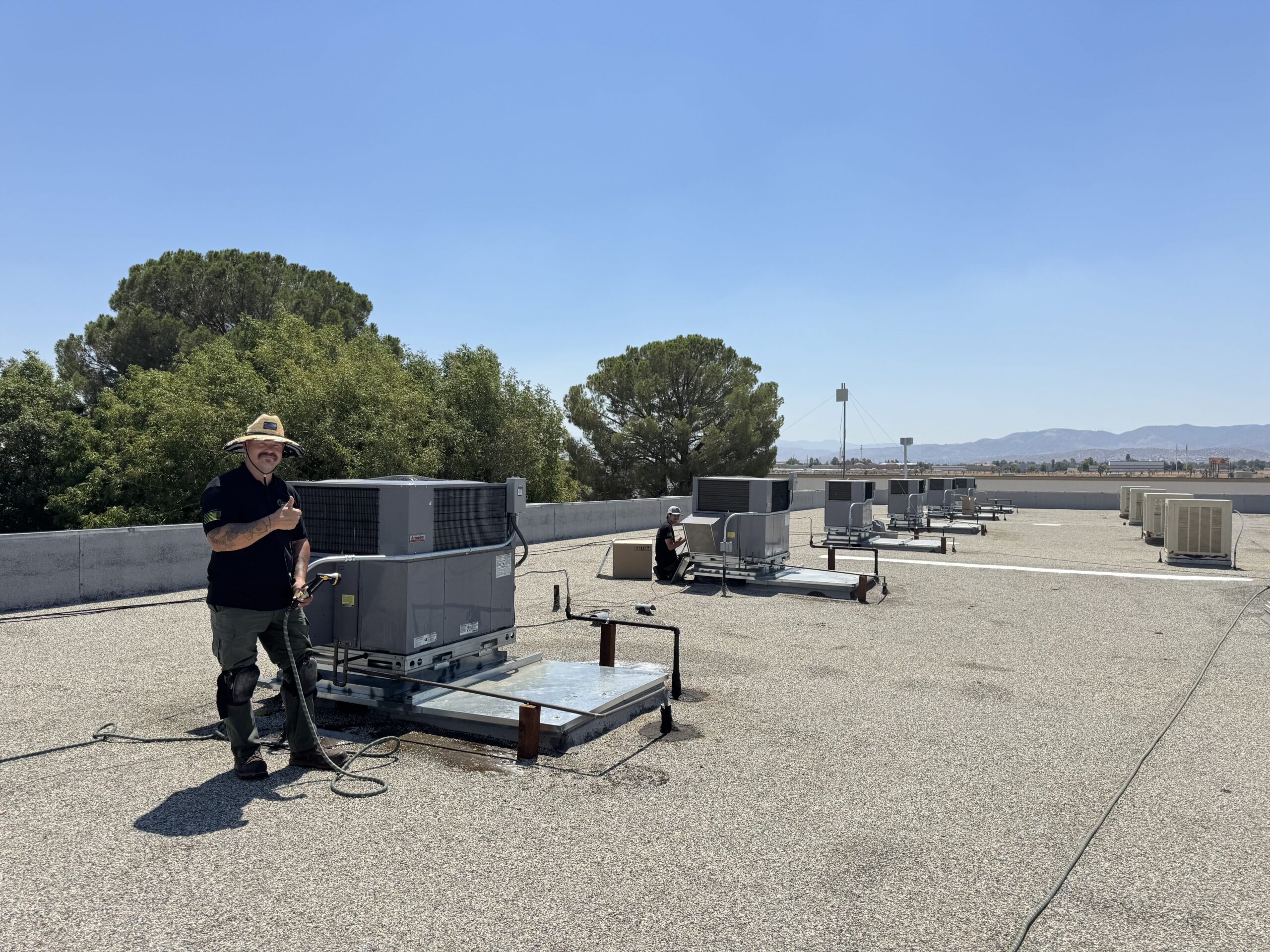 Two Origin Heating & Air technicians performing AC maintenance on multiple rooftop HVAC units at a commercial property in Palmdale, CA, with mountain views in the background