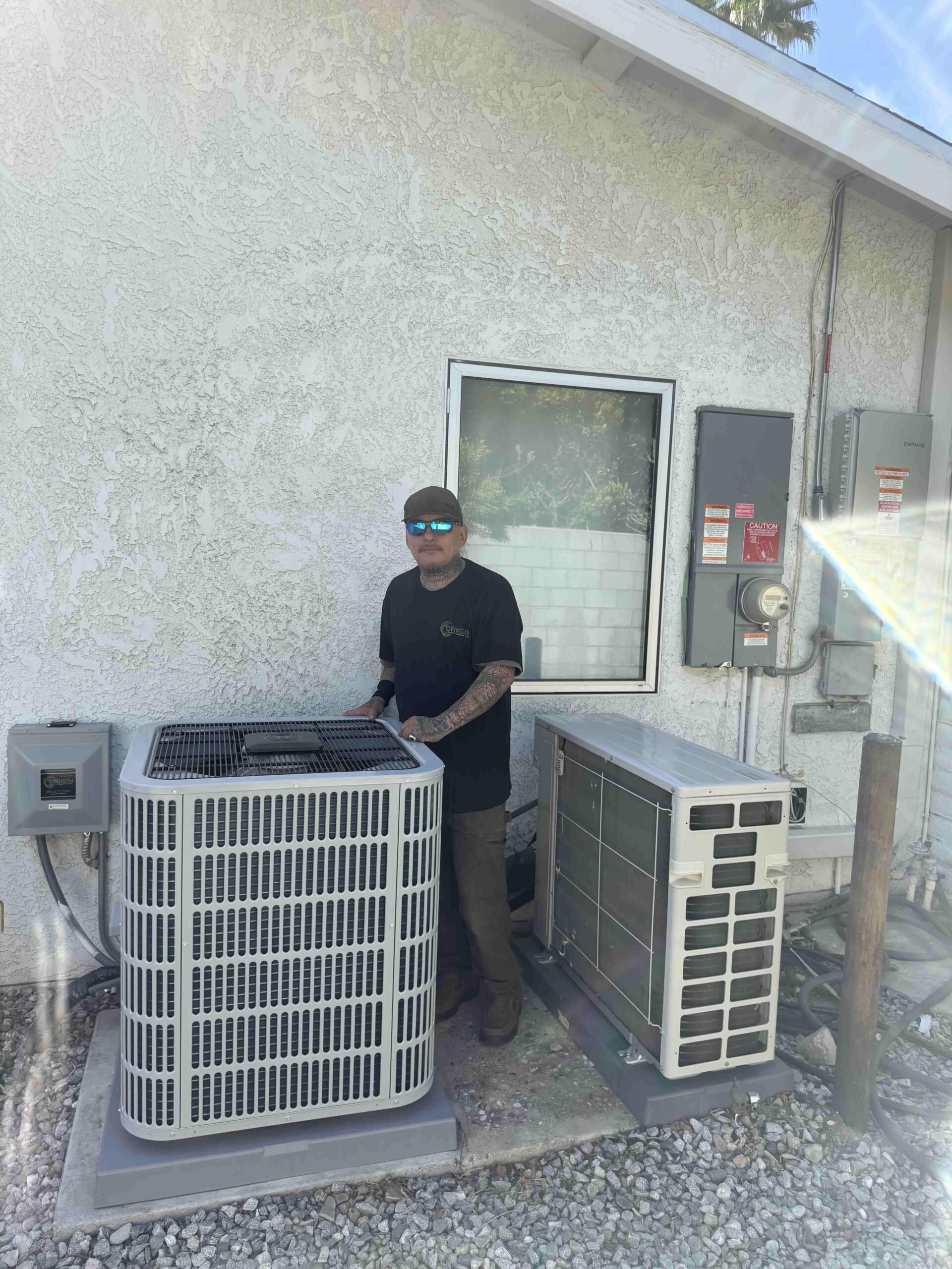 An Origin Heating & Air HVAC technician standing beside a residential AC condenser unit and mini-split outdoor unit against a stucco wall in Palmdale, ready for summer AC maintenance